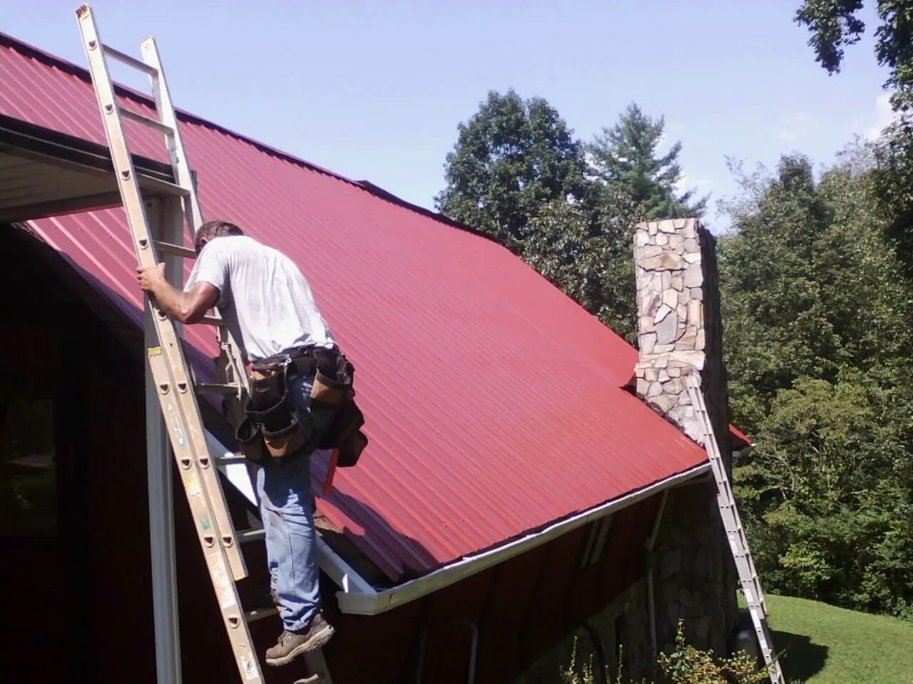 Roofer on a ladder beside a red metal roof and stone chimney — Soffit & Fascia Repair by Roofing Brothers in Falkner